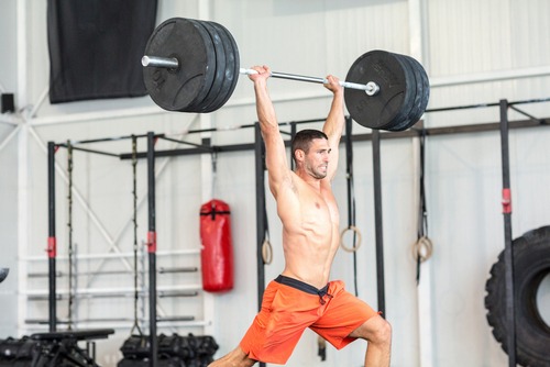 Shirtless man performing a barbell overhead press in a gym for strength training.