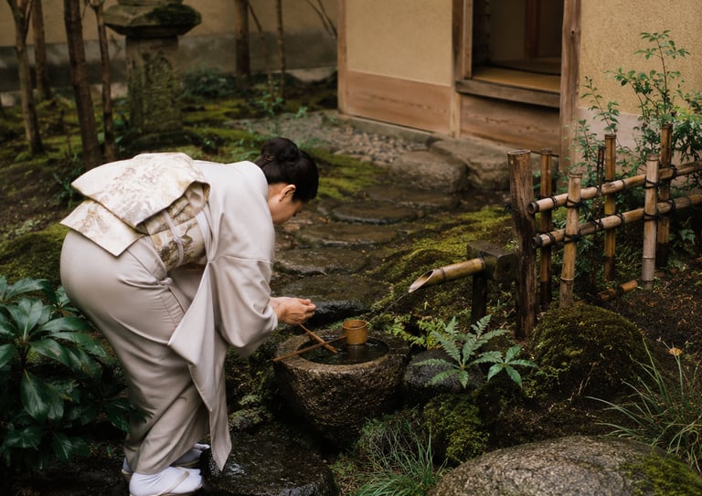 A woman in a traditional Japanese kimono performs a purification ritual at a stone water basin in a mossy garden.