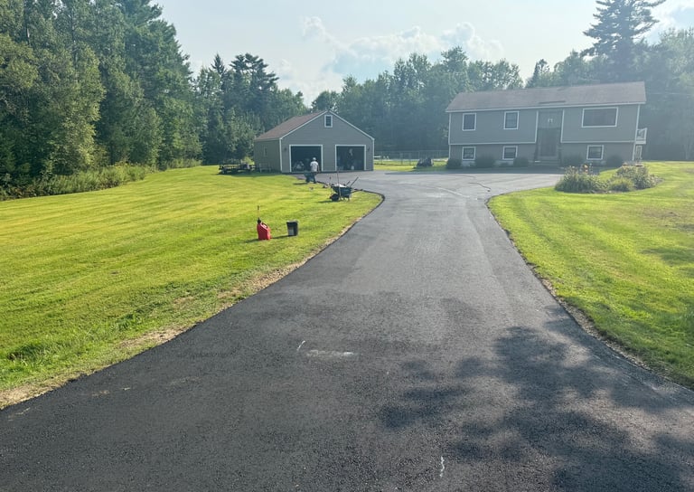 Freshly paved asphalt driveway leading to a residential house and detached garage with a green lawn.