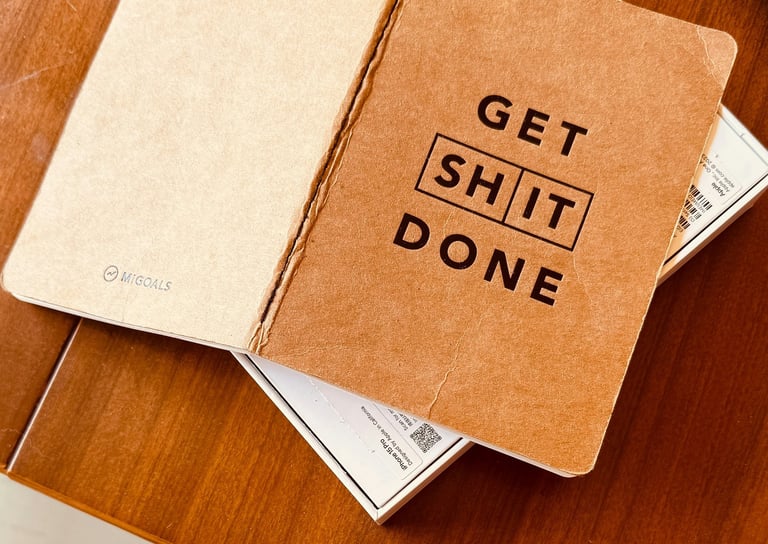 Open MiGoals brown kraft paper notebook with Get Shit Done motivational text on a wooden desk.