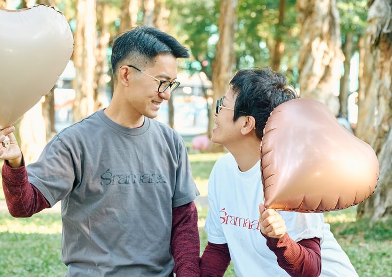 A smiling couple holds heart-shaped balloons while looking at each other in a sunny park.
