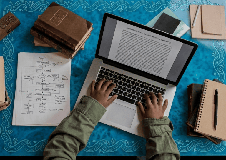 Person typing on a laptop for academic research with vintage books and flowcharts on a blue desk.