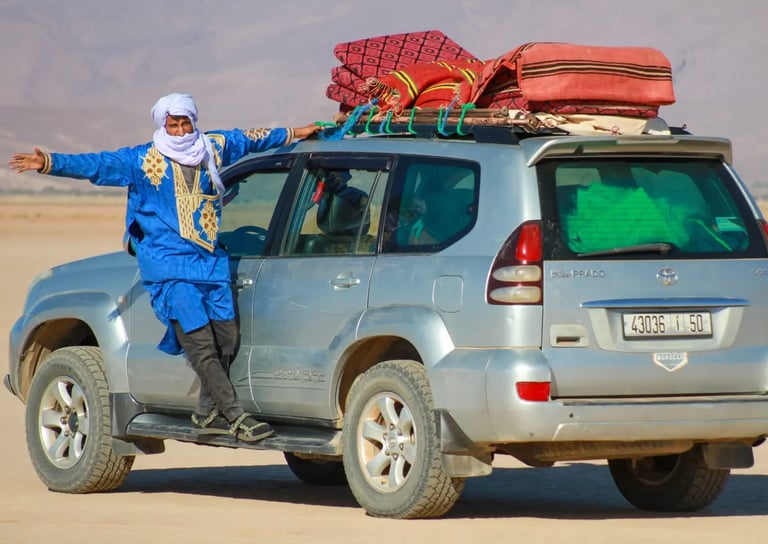 A man in traditional nomad dress stands on the running board of a 4x4 during a Moroccan Sahara trek