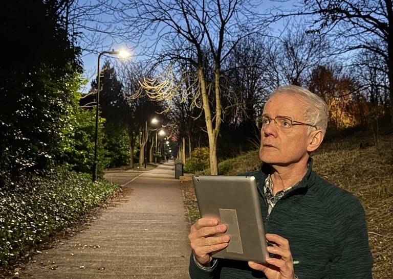 Photograph of a man, standing at the side of a footpath, at night, holding a tablet computer.