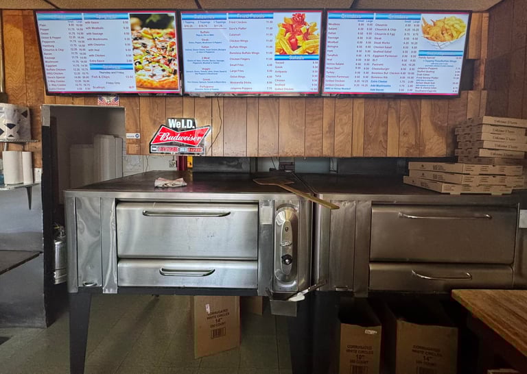Professional pizza oven in a restaurant kitchen below digital menu boards displaying pizza and grinder prices.