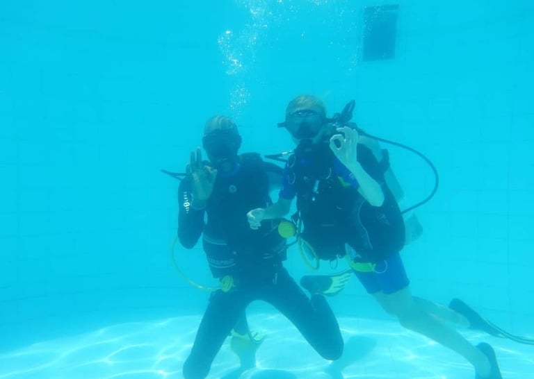 Two scuba divers give the OK sign while practicing underwater skills in a swimming pool.