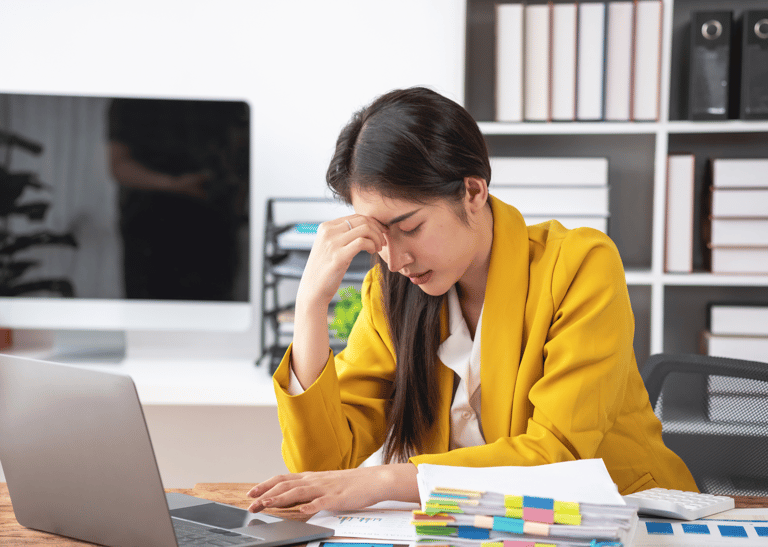 a woman in a yellow jacket is sitting at a desk with stressed face looking at laptop