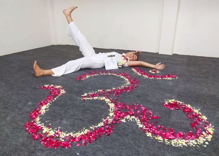 Eva lying on the ground next to a flower mandala, smiling happily while holding her Yoga Teacher Training diploma in Rishikes