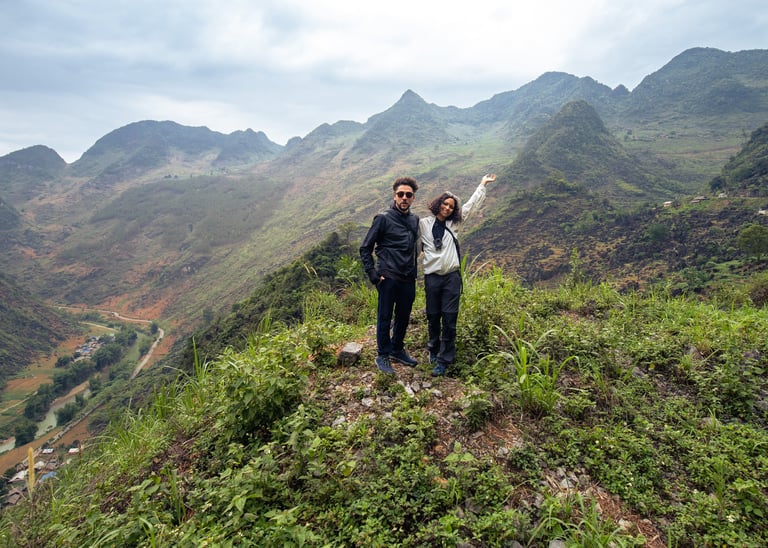 A couple standing on a grassy mountain edge posing for the camera. in the background are the beautiful Ha Giang mountains.