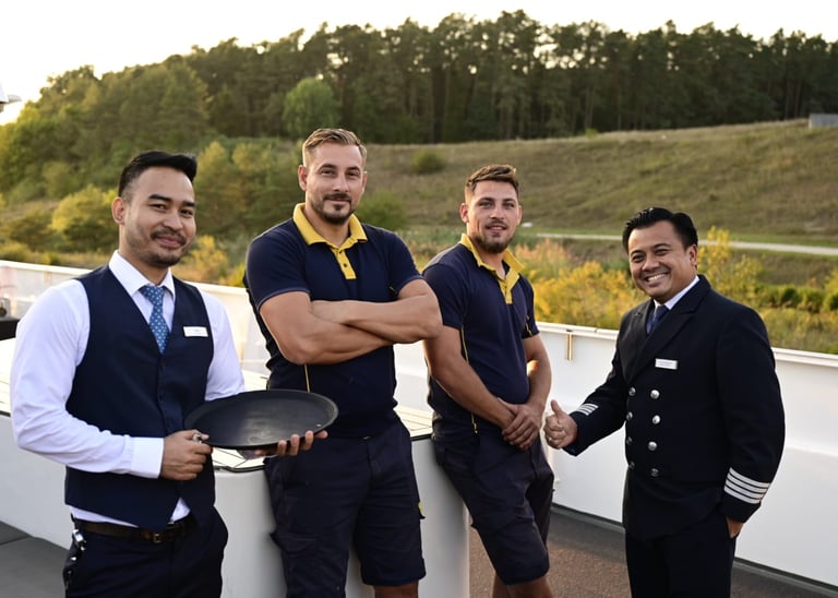 A smiling professional river cruise crew in uniform posing on a ship deck with a scenic green background.