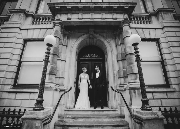 Black and white photo of bride & groom standing at a grand building entrance,captured by Fred Studio