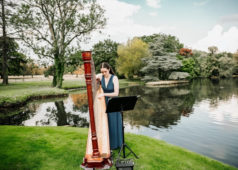 Harpist performing by a lakeside during an outdoor wedding.