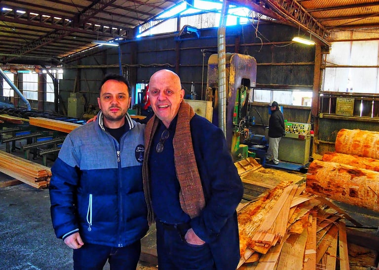 Two men standing inside a woodworking factory or sawmill surrounded by stacks of cut lumber and logs.