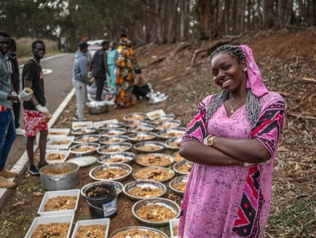 Una mujer afro con un vestido rosa parada frente a varios platos llenos de comida.