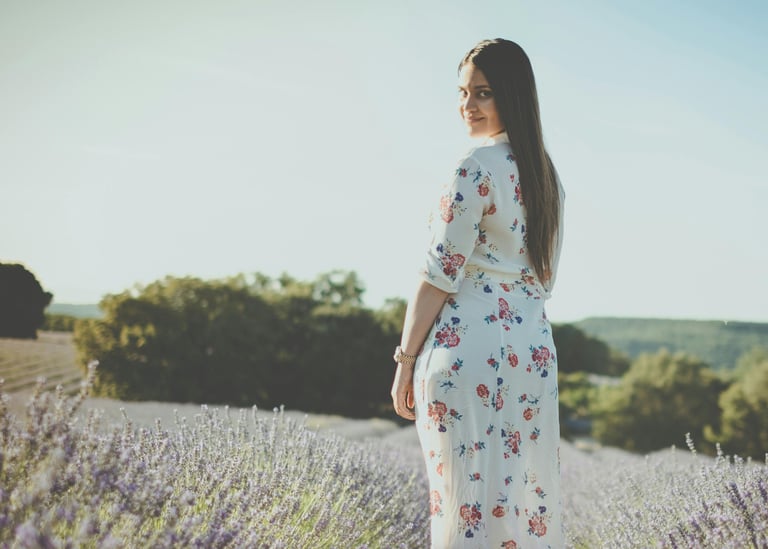 a woman in a white floral dress standing in a field of lavender. She looks at you and smiles.