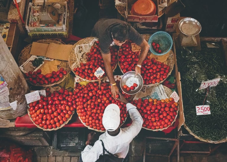 Overhead view of two men interacting at a market stall with baskets full of tomatoes, surrounded by various produce