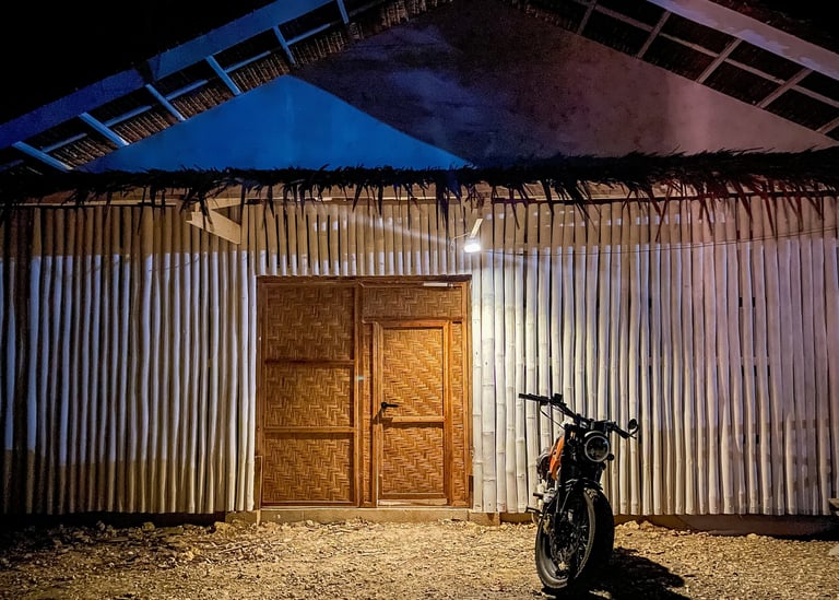 A vintage motorcycle parked at night in front of a rustic bamboo house with woven doors.