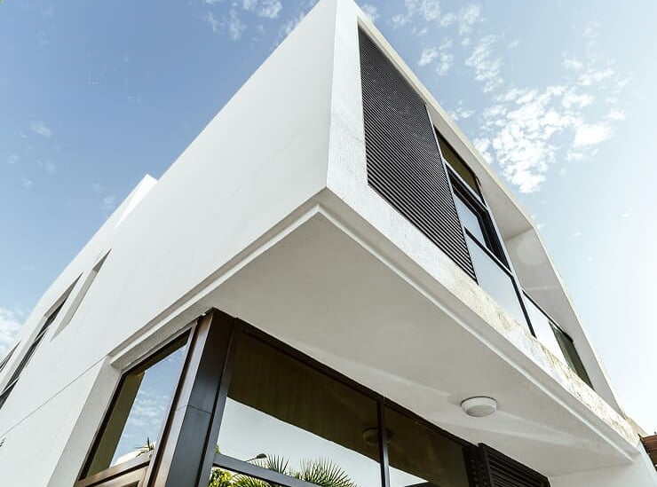 Low angle view of a modern white minimalist house with large glass windows and black shutters against a blue sky.