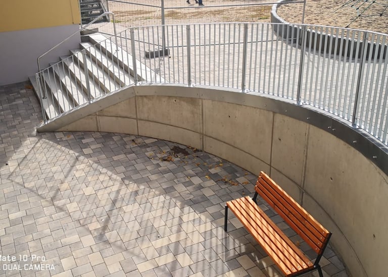 Modern outdoor courtyard with a wooden park bench, curved concrete wall, and metal railing stairs.