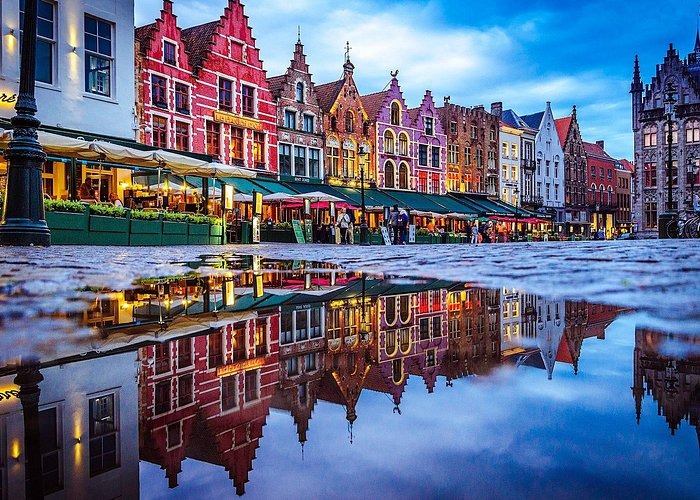 Colorful medieval architecture in Bruges reflected in a rainwater puddle at night.