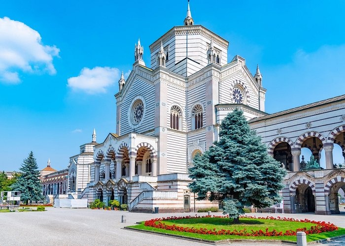 The grand Neo-Gothic facade of the Cimitero Monumentale in Milan under a bright blue sky.