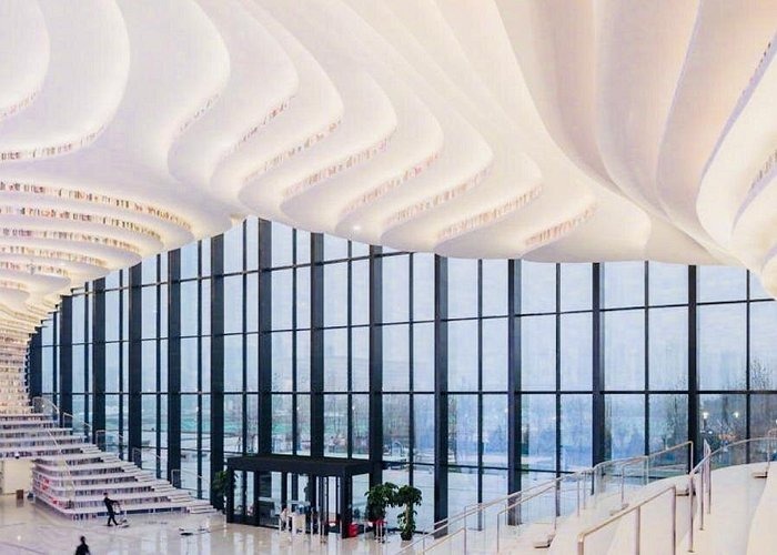 Modern library interior featuring white undulating terraced bookshelves and floor-to-ceiling glass walls.
