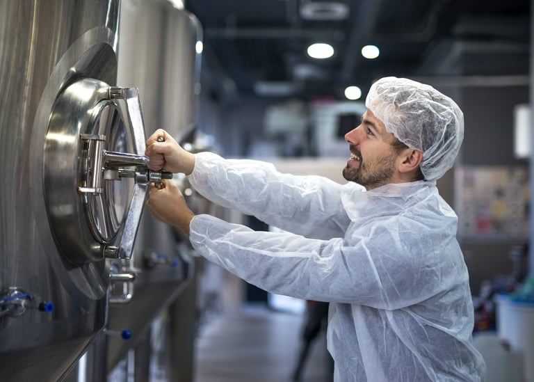 A brewery worker in protective gear adjusts a stainless steel fermentation tank.