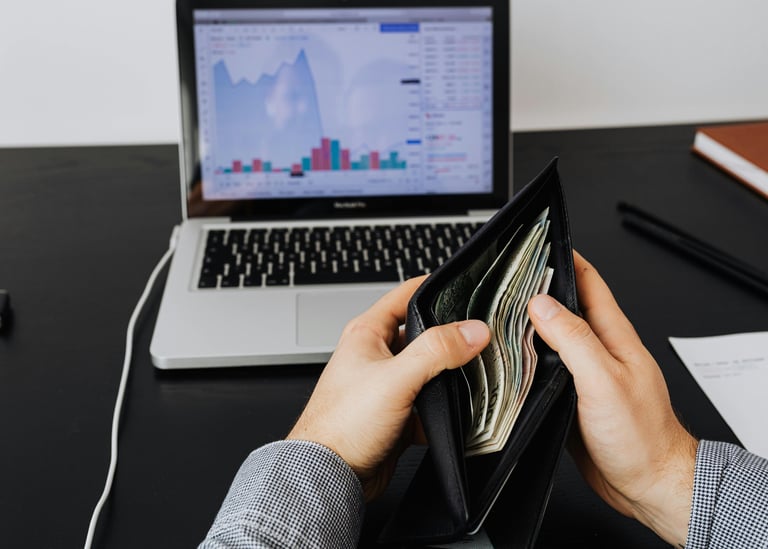 Notebook sobre uma mesa e duas mãos segurando uma carteira aberta com dinheiro dentro