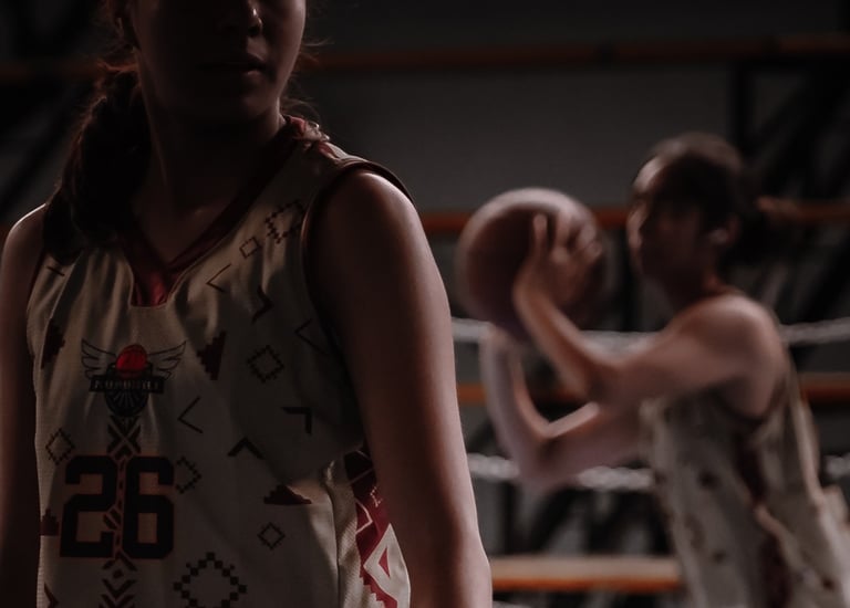 A female basketball player in a custom patterned jersey stands on court while a teammate shoots a ball.