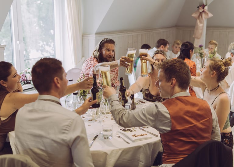Hochzeitsgäste stoßen fröhlich mit Biergläsern am Tisch an. Authentische Eventfotografie von Just Perspectives.