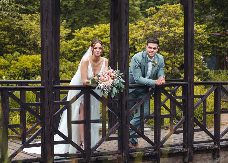 Brautpaar lehnt entspannt an einer Holzbrücke im Grünen. Hochzeitsportrait von Just Perspectives.