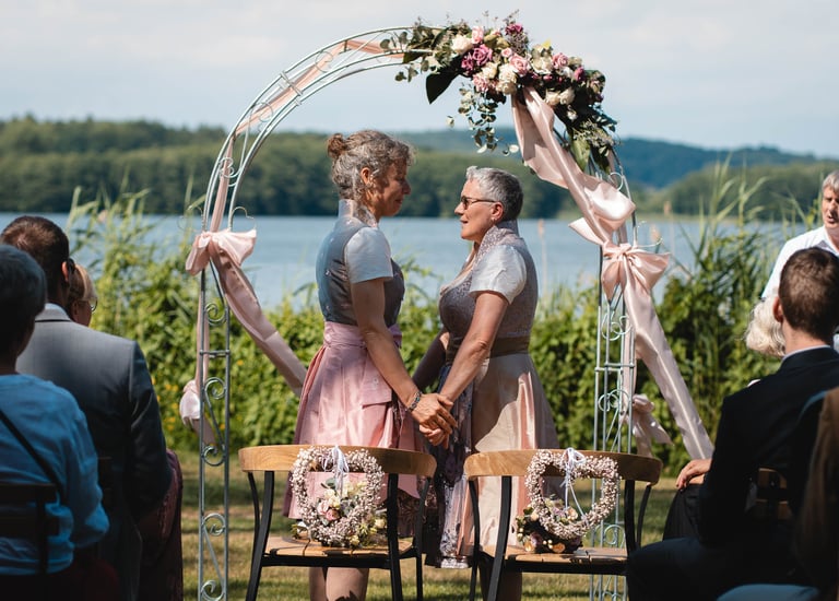 Zwei Bräute halten sich bei der freien Trauung unter einem Blumenbogen an den Händen. Foto: Joel Pingel.