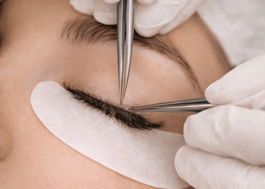 A beauty technician applies professional eyelash extensions using precision tweezers and under-eye pads.
