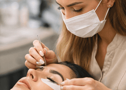 Professional lash artist applying eyelash extensions to a client using tweezers under a ring light.