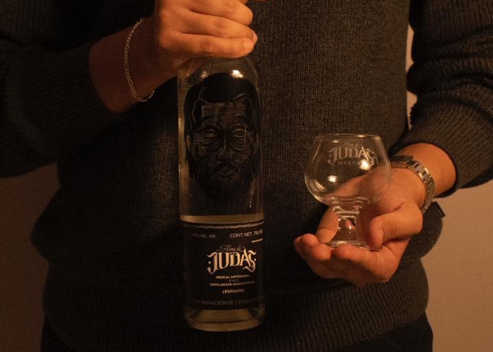 A person holding a bottle of Alma de Judas artisanal mezcal and a small tasting glass.