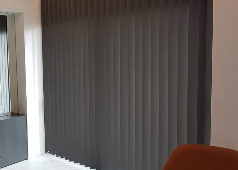 Modern charcoal grey vertical blinds installed on a large window with warm cove lighting.