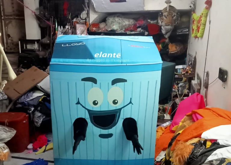 A person wearing a blue Lloyd Elante washing machine mascot costume in a workshop filled with fabrics.