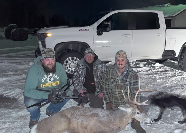 Three hunters posing in the snow with a harvested buck and thermal drone near a white truck.