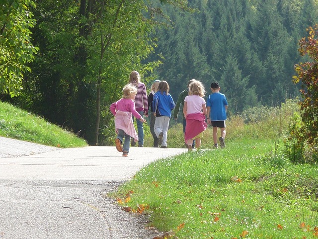 A group of happy children running and walking along a sunny paved path near a green forest.