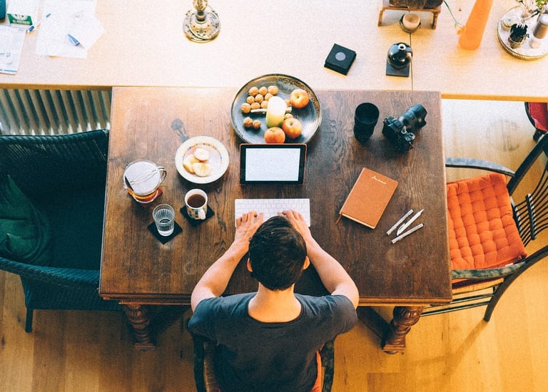 A top-down view of a writer working at a wooden table with a laptop, notebook, coffee, and snacks, surrounded by creative too