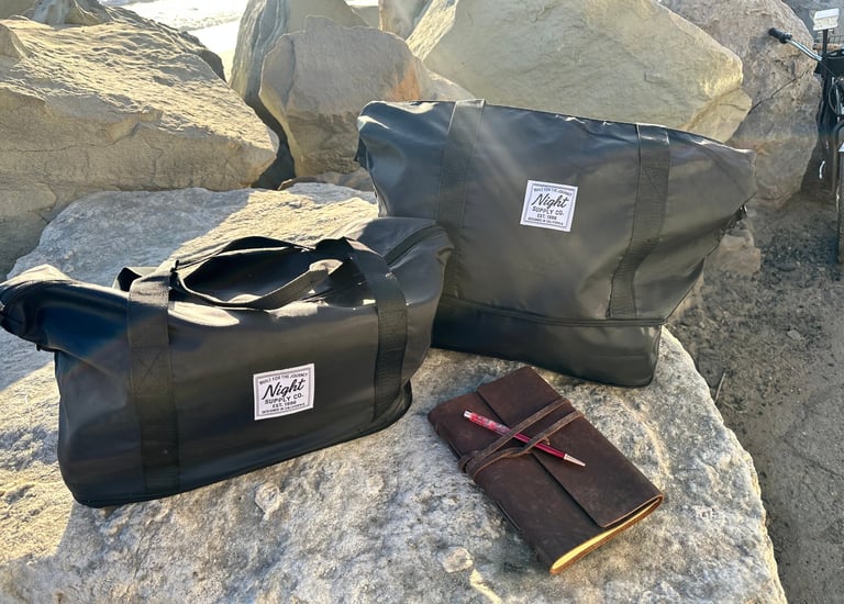 Two black Night Supply Co. duffel bags on rocks beside leather journal and pen.
