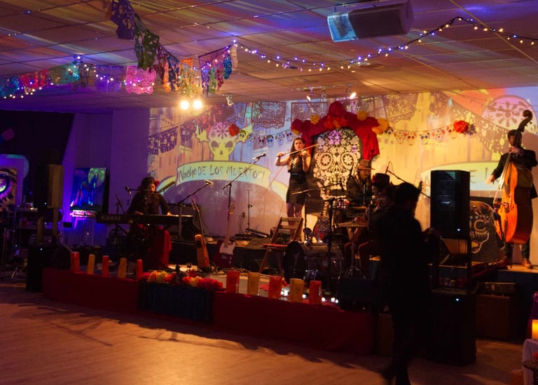 A live band performs on a stage decorated for Dia de los Muertos with colorful papel picado and string lights.