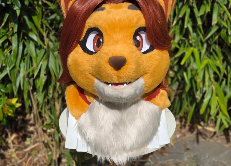 Custom orange feline fursuit head with striped ears and brown hair displayed outdoors.