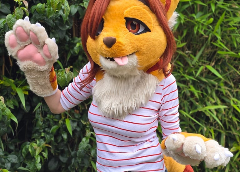 Person in a yellow and orange striped feline fursuit waving near green foliage.