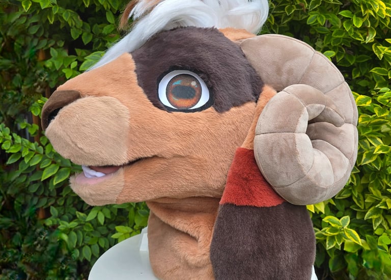 Custom brown ram fursuit head with curved horns and white hair displayed outdoors against green leaves.