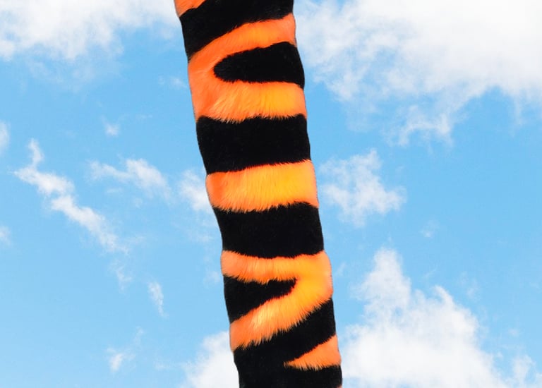 Handmade orange and black tiger faux fur tail for furry cosplay accessories with a white tip against a blue sky.