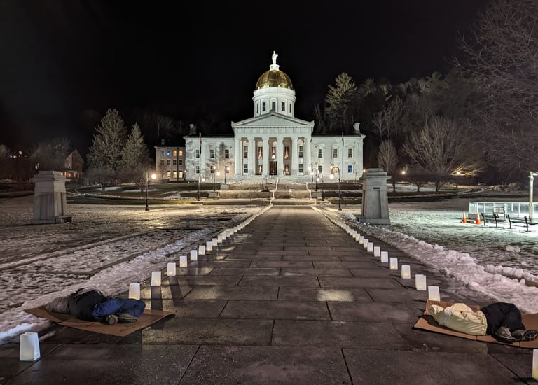 A nighttime picture of the lit Vermont capitol building. Two people are sleeping on the concrete.