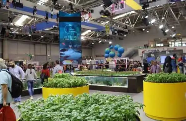 An indoor exhibition or trade show filled with people walking around various displays. Large planter
