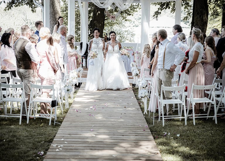 Dieses Farbfoto zeigt zwei Bräute, die in ihren Hochzeitskleidern einen Holzsteg entlanggehen, der mit verstreuten Blütenblät