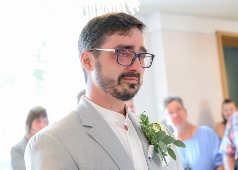 Emotionaler Bräutigam im hellgrauen Anzug mit Brille und Ansteckblume, der während der Trauung Tränen in den Augen hat.